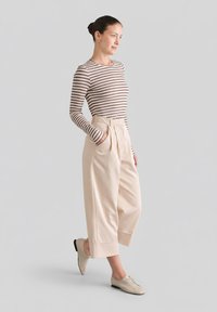 Kobieta w beżowych szerokich, krótkich spodniach, beżowych butach na sznurowadła oraz brązowo-białej, paskowanej bluzce z długim rękawem, stojąca z rękami w kieszeniach.
