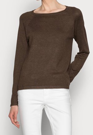 Femme portant un pull marron à manches longues avec un large décolleté et un pantalon blanc, debout avec une main dans le dos.