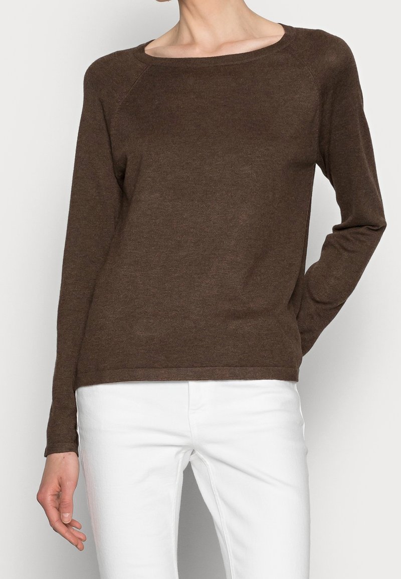 Femme portant un pull marron à manches longues avec un large décolleté et un pantalon blanc, debout avec une main dans le dos.