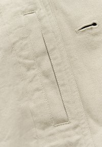 Jasnobeżowa tkanina o widocznej fakturze i ukośnych paskach. Posiada boczną kieszeń i małe wycięcie. Szwy są wyraźnie zszyte.