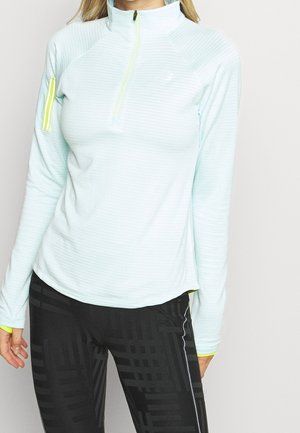 Žena nosící světle modrý sportovní top s dlouhým rukávem a žlutým zipem a černé legíny s vzorem, stojící před jednobarevným pozadím.