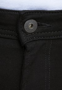 Los jeans de mezclilla negros presentan un cierre con botón de metal y detalles de costura visibles. La tela tiene una apariencia suave y texturizada.