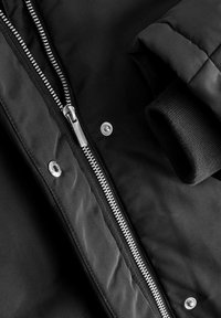 Giacca a vento nera con zip argentata, bottoni a pressione e polsini a coste. Tessuto morbido e liscio con una finitura opaca.