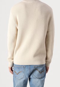 Pull beige côtelé à col montant, poignets ajustés et motif texturé, associé à un jean bleu clair avec des coutures courbes sur les poches.