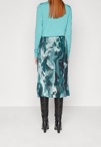 Pull turquoise et jupe mi-longue à motif abstrait multicolore. La jupe a une texture fluide et une fente sur le côté, associée à des bottes noires jusqu'aux genoux.