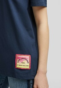 T-paita laivastonsinisenä, lyhythihaisena, jossa on teksturoitu läppä vaaleanpunaisena, keltaisena ja mustana, esittäen brändin nimeä ja osoitetta Pariisissa.