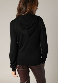 Zwarte, hoodie trui met een gestructureerde brei. Heeft een losse pasvorm en geribbelde manchetten. Gepaard met glanzende bordeauxrode broek.