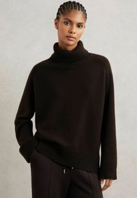 Suéter de punto marrón oscuro con un cuello alto de canalé. Presenta hombros caídos, puños amplios de canalé y un corte relajado. Textura suave.