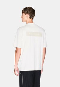 T-shirt bianco in cotone con maniche corte, caratterizzata da un sottile rettangolo logo beige sul retro. Tessuto liscio, vestibilità rilassata e design semplice.