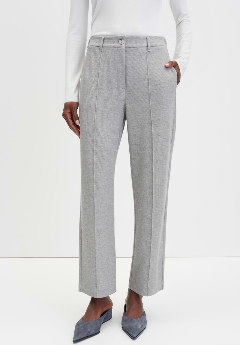 Personne portant un pantalon large gris clair sur mesure avec pli sur le devant, un haut blanc à manches longues et des talons gris à bout pointu, debout avec la main dans la poche.