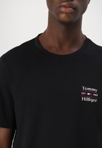 Zbliżenie na osobę w czarnej koszulce z małym białym i czerwonym logo Tommy Hilfiger oraz tekstem po lewej stronie klatki piersiowej.