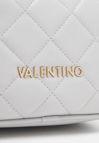 Fehér steppelt kézi táska, amely kiemelkedő arany "Valentino" logóval, sima textúrával és díszes gyémántmintás varrással rendelkezik.