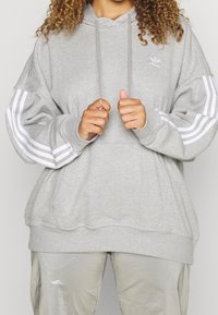 Sweatshirt à capuche gris avec des accents blancs à trois rayures sur les manches. Présente une coupe décontractée, une capuche avec cordon de serrage et des poignets côtelés.