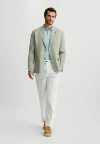 Blazer de lino verde claro sobre una camisa de botones azul claro, combinado con pantalones blancos y zapatos beige, creando un conjunto informal.