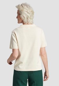 Pull à manches courtes crème avec col et poignets côtelés, associé à un pantalon vert, mettant en avant une coupe décontractée et un tissu doux et texturé.