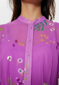 Bluză violet din material transparent, cu guler mandarin, nasturi frontali și modele florale cu accente metalice. Textură netedă, material ușor.
