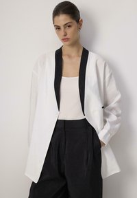 White, lightweight kimono-style jacket with black collar, long sleeves, and a loose fit, paired with black pleated pants.