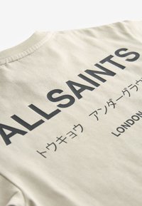 Bézs póló nagy fekete "ALLSAINTS" felirattal, kisebb japán karakterekkel és az alján hátul nyomtatott "LONDON" felirattal.