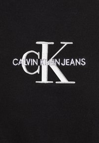 Tejido negro con un destacado logotipo bordado, "CALVIN KLEIN JEANS," en blanco y acentos plateados, con un diseño de fuente limpio y moderno.