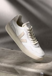 Sneaker bianco con tomaia in mesh testurizzato e accenti crema, dotato di suola liscia e dettagli beige sui lati e sulla linguetta.