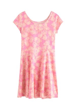 Smilende pige står iført en kortærmet lyserød blomsterkjole og lysefarvede blondeballetsko mod en ensfarvet hvid baggrund.