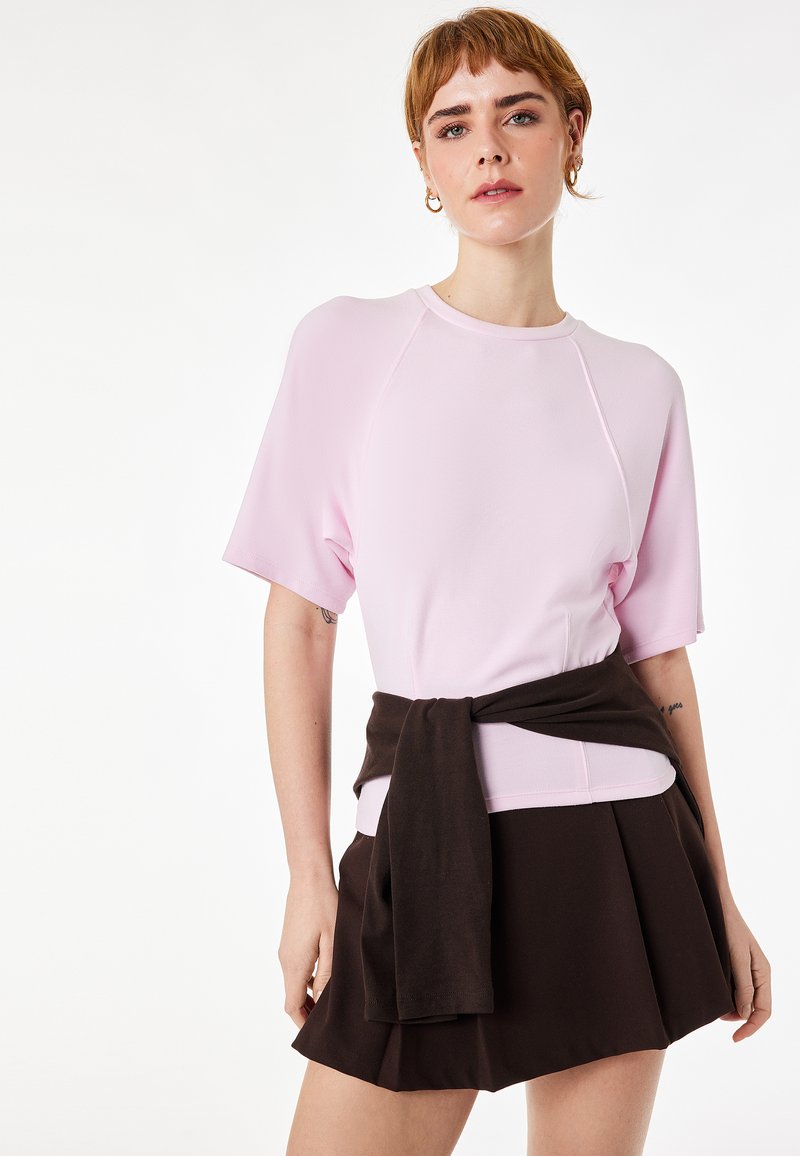 Jeune femme aux cheveux courts portant un haut à manches courtes rose clair et une jupe noire avec un pull noir noué autour de la taille.