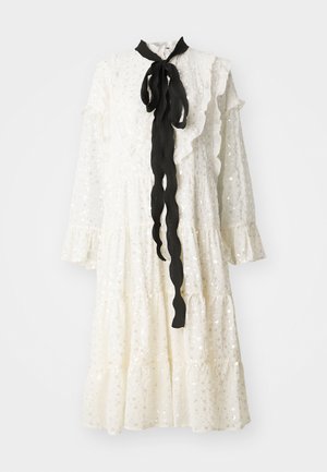 Valkoinen mekko, jossa on röyhelökaulus ja hihat, mustalla nauhaketjulla ja kankaan pinnassa yksityiskohtia strasseilla. Kerroksellinen muotoilu, jossa on virtaava siluetti.