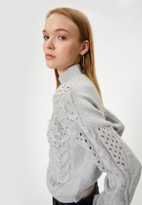 Pull en maille gris clair avec un col montant, motif en fausse-torse, et clous dorés décoratifs le long des épaules et des bras.