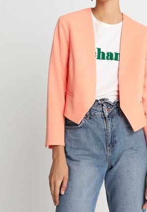 Vrouw draagt een cropped perzikkleurige blazer over een wit grafisch t-shirt, ingestopt in hooggetailleerde blauwe jeans met paperbag taille.