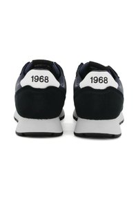 Un paio di sneakers nere e bianche viste da dietro, ciascuna con "1968" stampato sulla linguetta del tallone sopra le suole bianche e le suole nere.