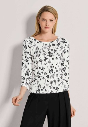 Frau trägt ein weißes Langarmshirt mit schwarzen Blumen- und Schleifenmustern, kombiniert mit schwarzen Hosen, und steht vor einem schlichten Hintergrund.