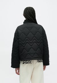 Czarna pikowana kurtka z falistym wzorem, z wysokim kołnierzem i opuszczonymi ramionami. Tył ma gładką teksturę.