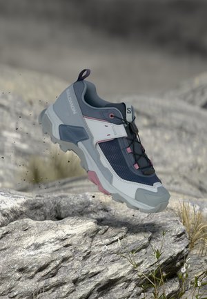 Zapatilla de trail running en gris, azul marino y rosa en el aire sobre un terreno rocoso con pequeñas plantas y un fondo natural desenfocado.