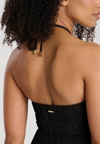 Kvinna med lockigt hår iklädd en svart strapless klänning med halterneck och guld accenter, visad från baksidan och sidan.