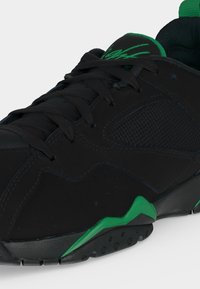 Μαύρο αθλητικό παπούτσι με πράσινες λεπτομέρειες, υφασμάτινα πάνελ και μαύρα κορδόνια, εμφανιζόμενο σε κοντινή πλευρική όψη.
