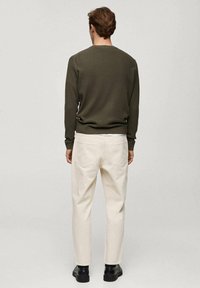 Uomo con capelli castani corti che indossa un maglione verde scuro, pantaloni avorio e scarpe nere, in piedi di spalle contro uno sfondo grigio.