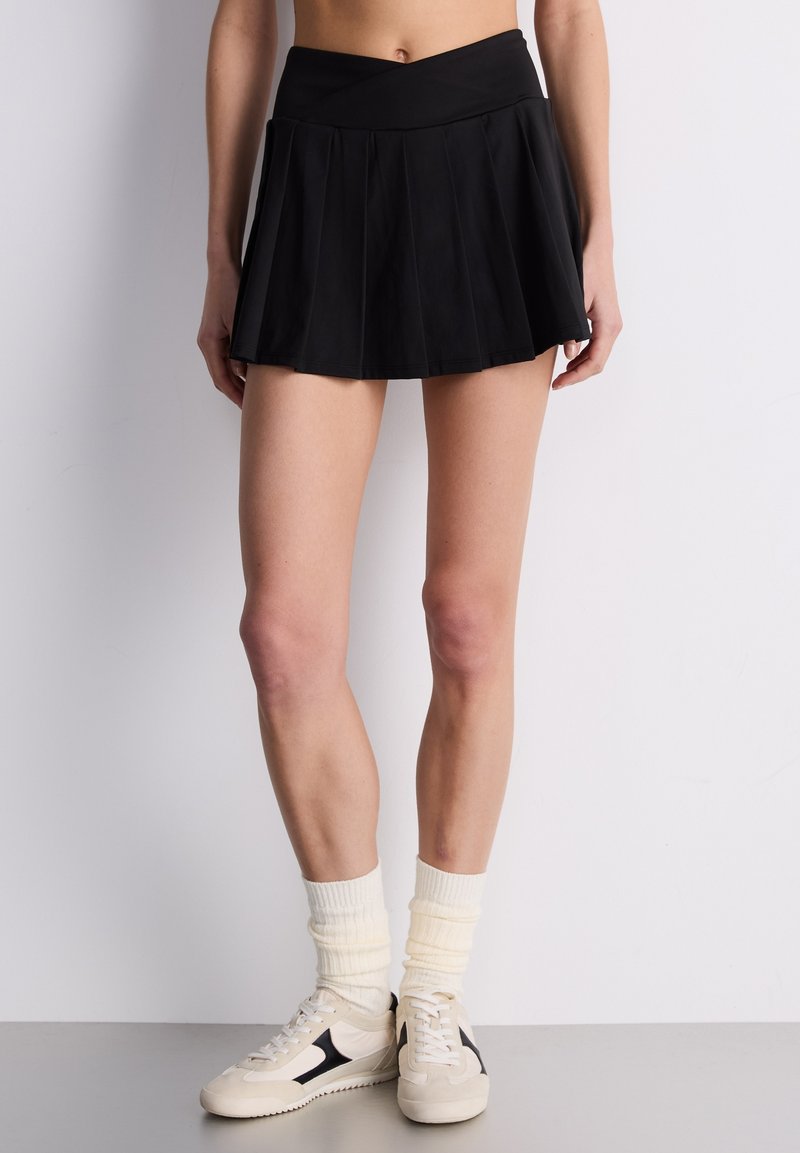 Czarna plisowana spódnica z dopasowanym pasem, wykonana z gładkiej tkaniny. Połączona z jasnymi skarpetkami i casualowymi trampkami.