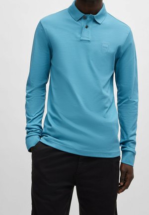 Mężczyzna ubrany w jasnoniebieską koszulkę polo z długim rękawem i dwoma guzikami oraz czarne spodnie, stojący z prawą ręką częściowo w kieszeni.