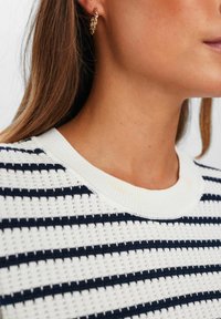 Balts adīts džemperis ar tumši zilu horizontālu svītru, ar ribotu apaļu kakla izgriezumu. Modelis valkā zelta ķēdes auskarus.