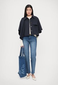 Chaqueta capa corta azul marino con botones, top beige, vaqueros azul claro y zapatos blancos. Lleva un bolso tote azul con un logo.