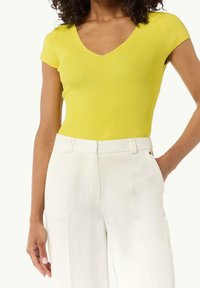 Gelb gerippter Kurzarm-Body mit V-Ausschnitt, kombiniert mit weißen, taillierten Hosen, die eine glatte Textur und einen körpernahen Schnitt aufweisen.