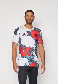 Miesten urheilupaita, jossa on valkoinen pohja ja sininen sekä punainen abstrakti kuvio, pyöreä kaula-aukko ja Adidas-logo rinnassa.