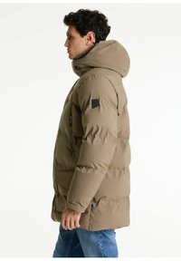 Peščeni puffer jopič s kapuco, ki ima zadrgo na sprednjem žepu, črne detajle in prešito obliko. Obučen čez modre kavbojke.