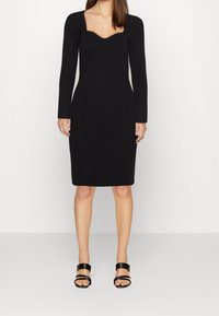 Femme portant une robe en maille noire moulante à manches longues avec un décolleté en cœur et des sandales à talons ouvertes noires, sur un fond uni.