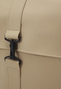 Sac impermeable beige avec finition mate, doté d'une boucle noire et du logo "RAINS" en relief. Texture lisse et détails de couture subtils.