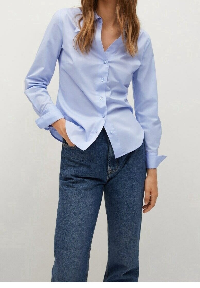 Femme avec une chemise bleu clair boutonnée et un jean bleu foncé, une main dans la poche, debout devant un fond clair uni.