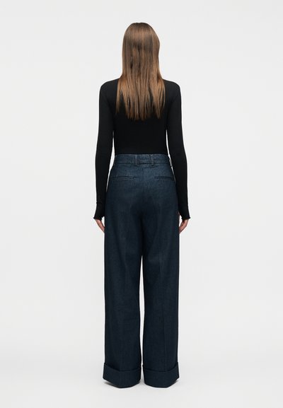 Γυναίκα με μακριά καστανά μαλλιά, που φοράει μαύρο μπλουζάκι με μακριά μανίκια και φαρδιές σκούρες μπλε παντελόνες, στέκεται με την πλάτη προς τα πίσω σε μια απλή φόντο.