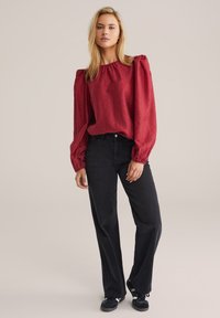 Blusa rossa con texture e maniche a sbuffo, abbinata a jeans neri a vita alta e sneakers scure. Design semplice e pulito.