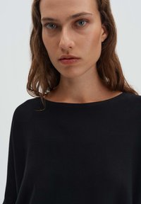 Jeune femme aux cheveux bruns ondulés et aux yeux bleus, portant un haut noir à encolure large, sur un fond gris clair uni.