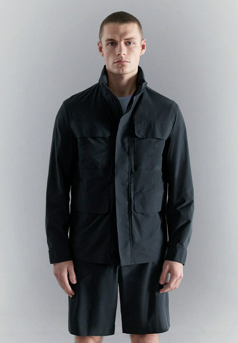 Jeune homme aux cheveux courts portant une veste noire à boutons avec quatre poches et un short noir assorti, sur un fond gris uni.